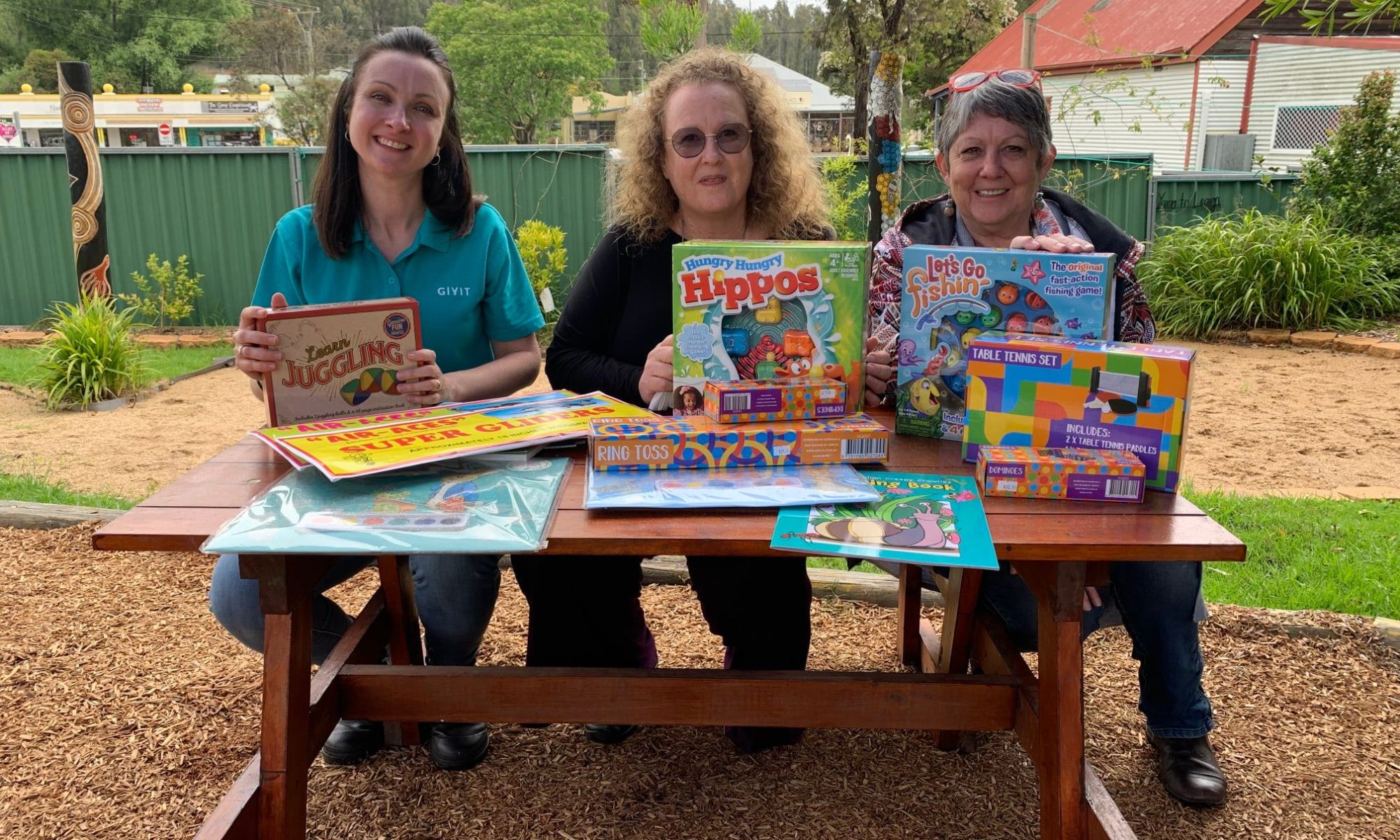 Three women sitting at a picnic table displaying a vairety of children's games, books and toys.
